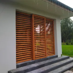 Trzy przesuwne, drewniane żaluzje w kolorze miodowym, zamontowane na zewnątrz budynku z szarymi schodami wejściowymi, widoczne elementy otoczenia.