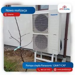 Zewnętrzna jednostka pompy ciepła Panasonic 12kW T-CAP zamontowana na metalowym stelażu przy ścianie budynku, obok małe drzewko z niebieską etykietą.