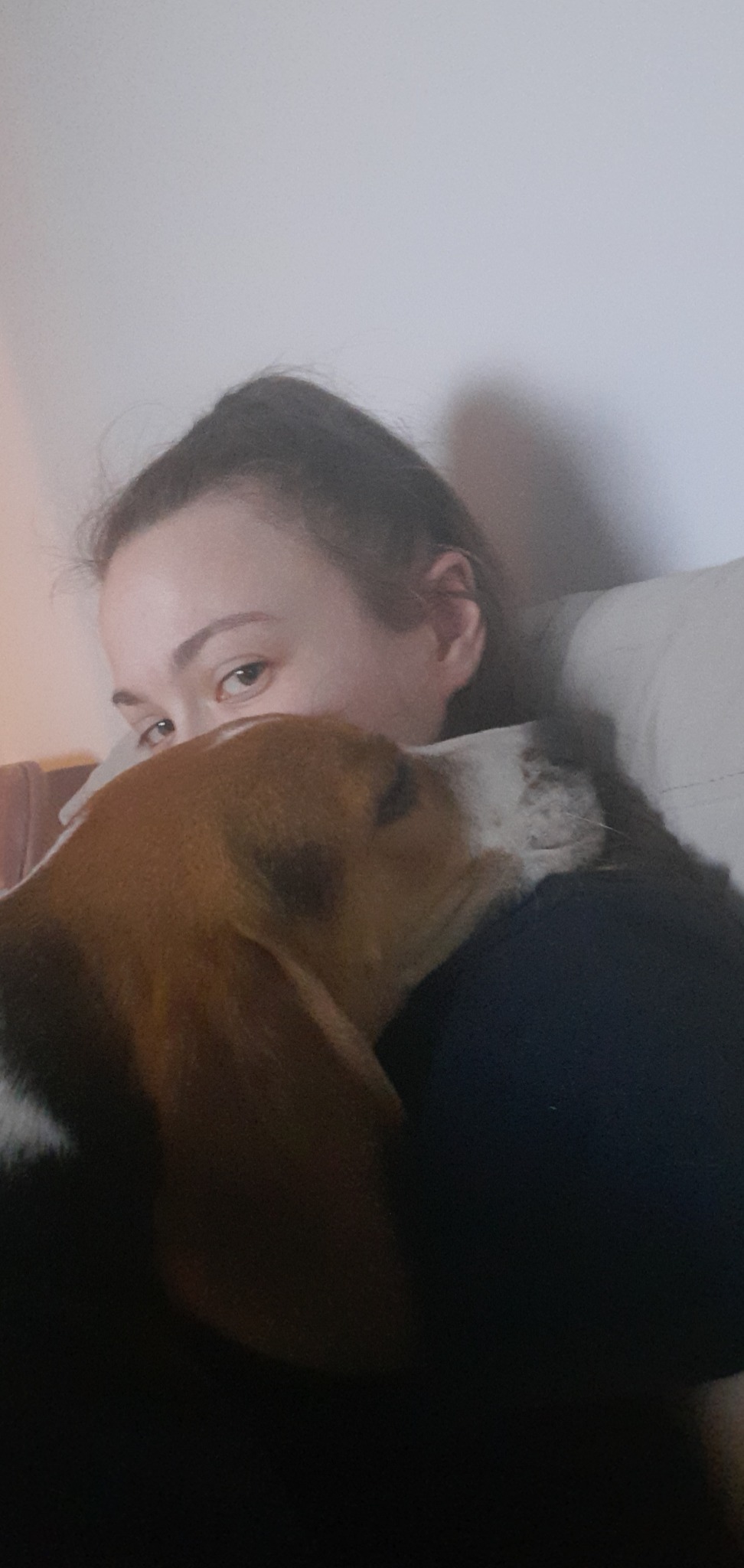 Młoda kobieta przytula psa rasy beagle, widoczne jej oczy i część twarzy psa, który opiera się o jej ramię na tle białej ściany.