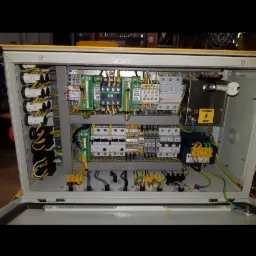 Otwarta szafa sterownicza z widocznymi elementami elektrycznymi, w tym zielone i żółte złączki, bezpieczniki oraz okablowanie. Tabliczka ostrzegawcza 500V.