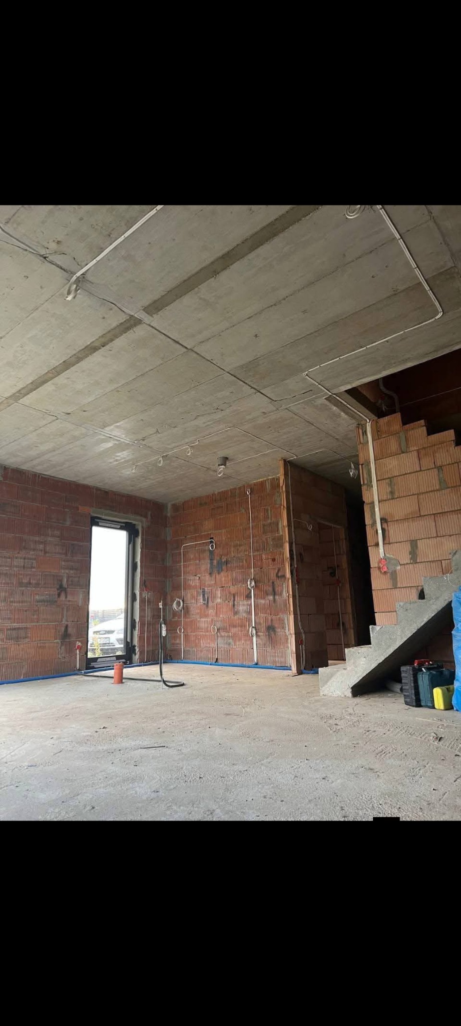 Surowy stan wnętrza z ceglanymi ścianami, widoczne instalacje elektryczne na ścianach i suficie, okno w tle, przygotowane do dalszych prac wykończeniowych.