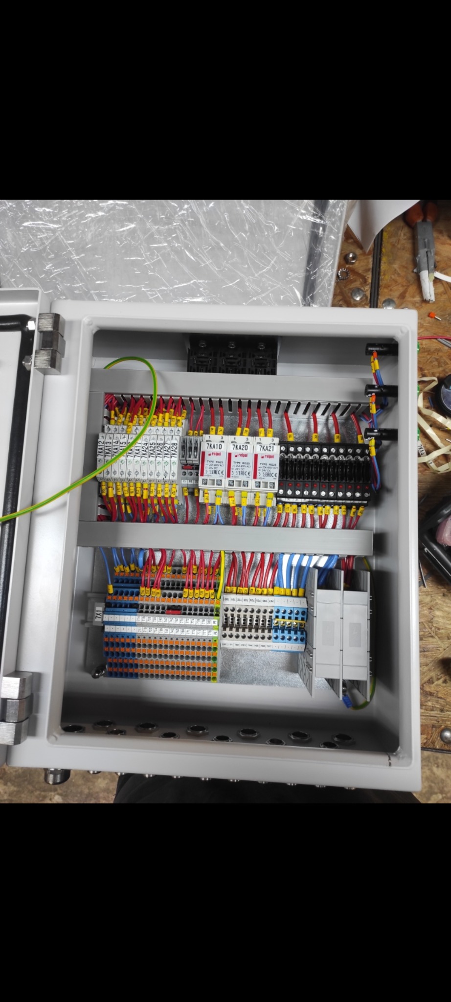 Rozdzielnica elektryczna w szarej obudowie z widocznymi przewodami, bezpiecznikami i elementami sterującymi. Przewody w kolorach: czerwonym, niebieskim, żółtym i zielonym.