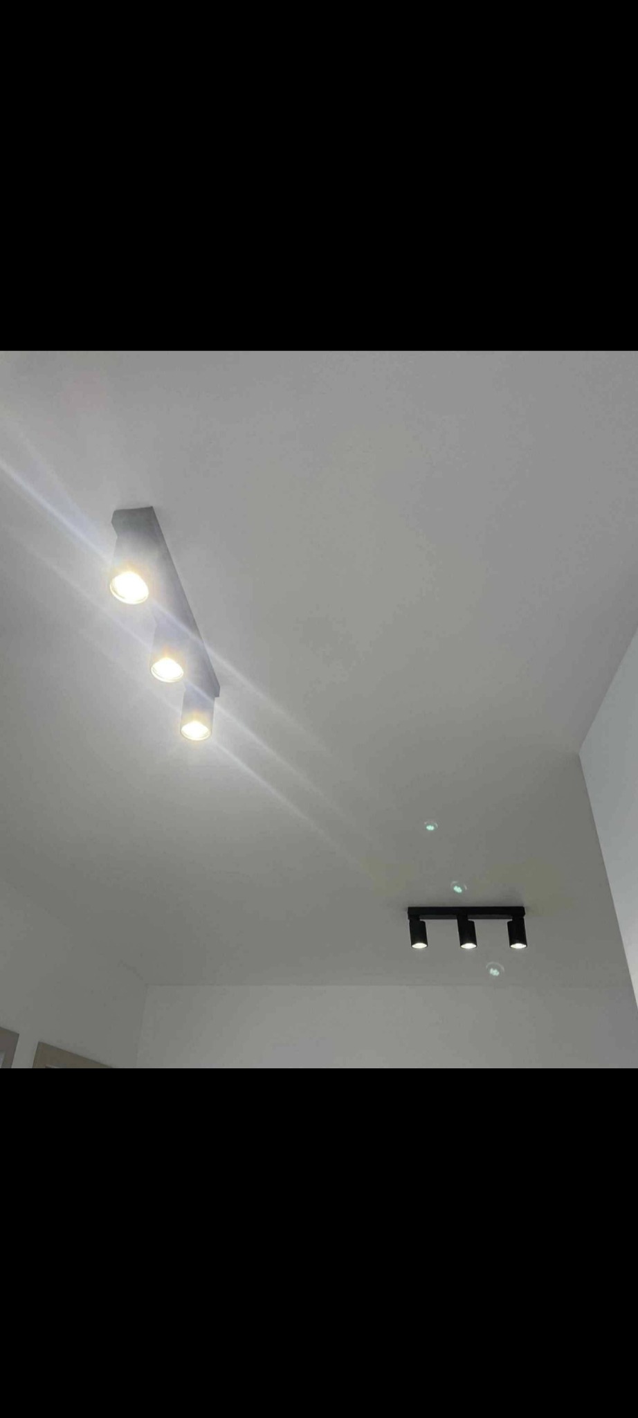 Dwa czarne reflektory sufitowe z trzema punktami świetlnymi każdy, skierowane w dół, oświetlają białe wnętrze. Widok z dołu, z lekkim kątem.