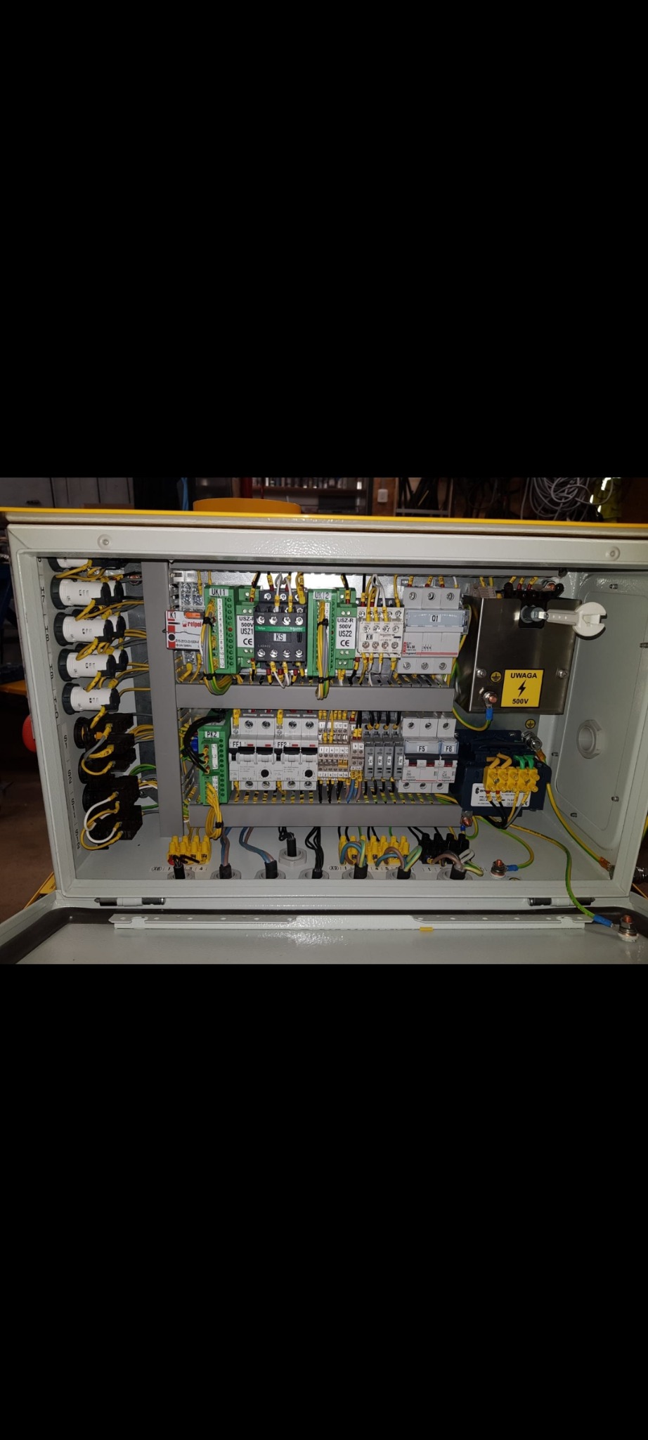 Otwarta szafa sterownicza z widocznymi elementami elektrycznymi, w tym zielone i żółte złączki, bezpieczniki oraz okablowanie. Tabliczka ostrzegawcza 500V.