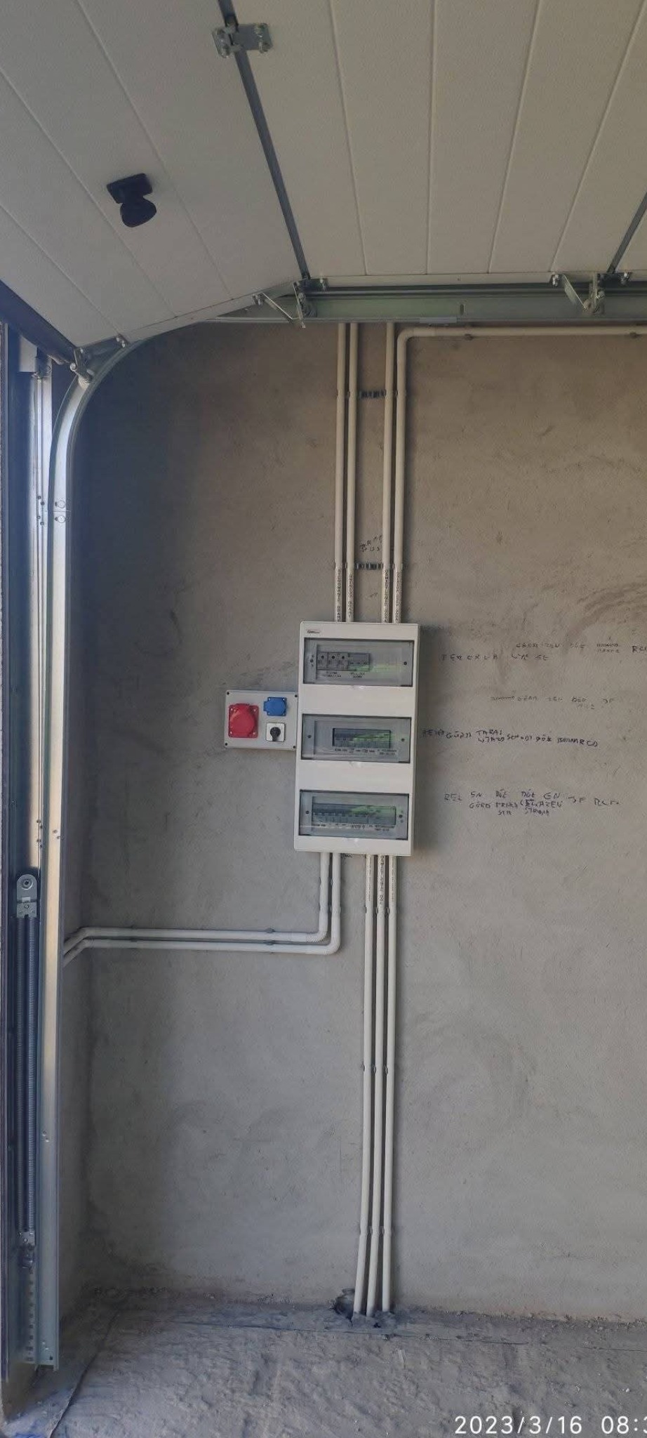 Rozdzielnia elektryczna z przewodami w białych rurkach na szarej ścianie w garażu z otwartą bramą segmentową. Widoczne gniazda siłowe.
