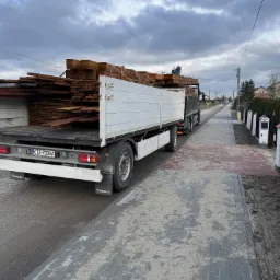 Ciężarówka z otwartą skrzynią ładunkową wypełnioną brązowymi deskami, zaparkowana na ulicy w pochmurny dzień.