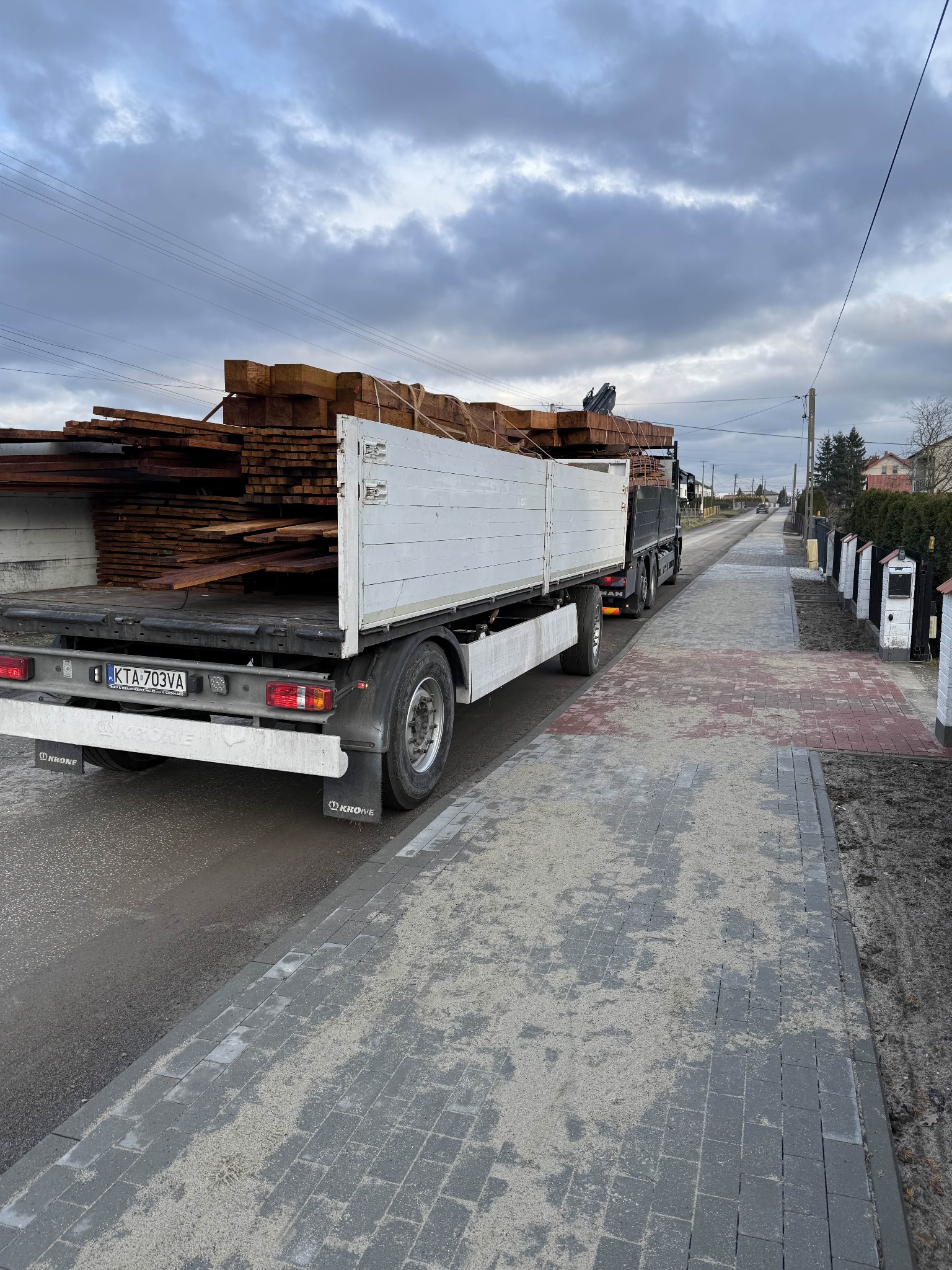 Ciężarówka z otwartą skrzynią ładunkową wypełnioną brązowymi deskami, zaparkowana na ulicy w pochmurny dzień.