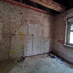 Wnętrze w trakcie remontu z odsłoniętą cegłą, bruzdami pod instalację elektryczną, przewodami wystającymi ze ściany i rurami na podłodze.