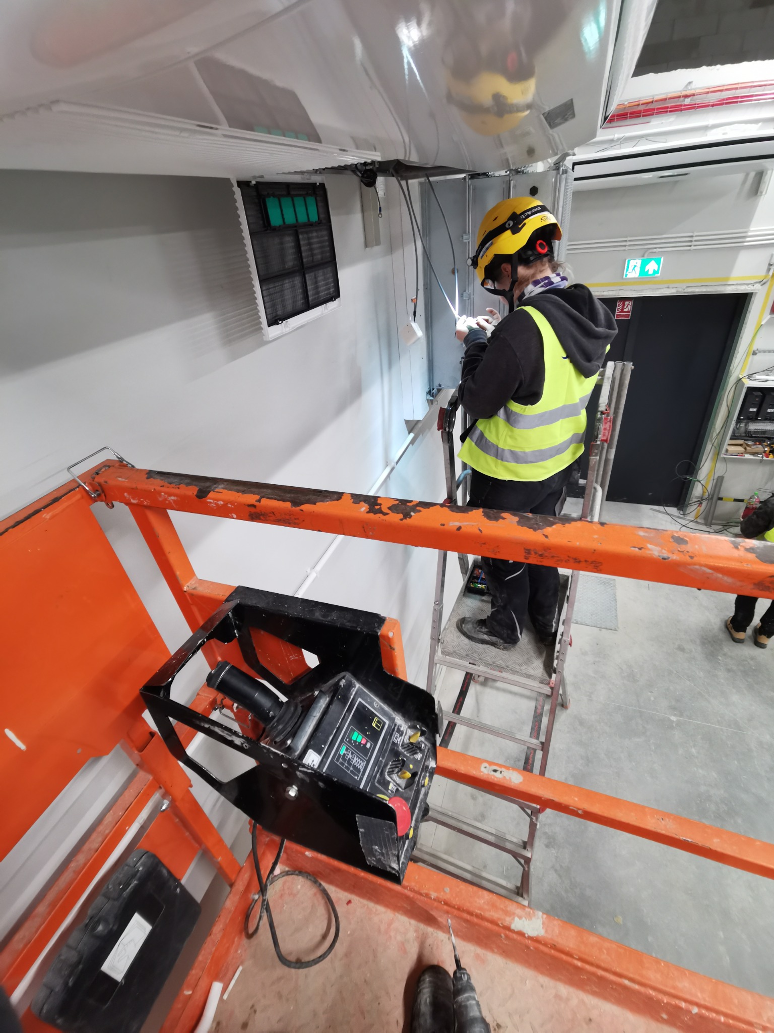Elektryk w kasku i kamizelce na podnośniku montuje instalację pod sufitem w hali. Widoczne przewody, kratka wentylacyjna i panel sterowania podnośnika.