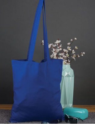 Niebieska torba bawełniana z uchwytami na tle szarej ściany, obok wazon z białymi kwiatami i okulary przeciwsłoneczne.