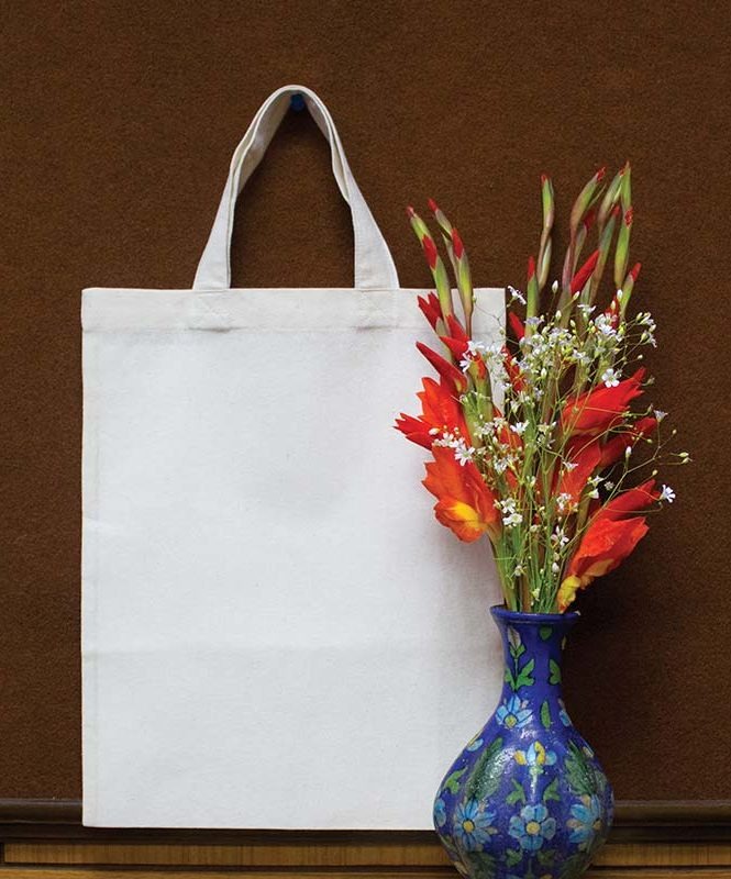 Biała, pusta torba bawełniana na zakupy, obok wazon z czerwono-białymi kwiatami na brązowym tle.