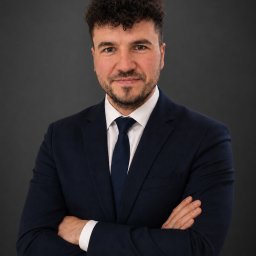 Kancelaria Adwokacka Daniel Graliński - Portret mężczyzny w ciemnym garniturze i krawacie, z rękami skrzyżowanymi na piersiach, na ciemnoszarym tle. Uśmiechnięty, z krótką brodą i kręconymi włosami.