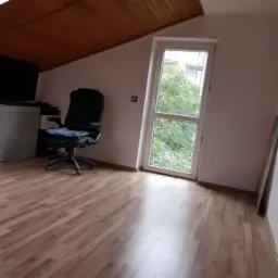 Jasny pokój na poddaszu z ukośnym sufitem, wykończony panelami podłogowymi w odcieniach brązu, widoczne krzesło biurowe, okno balkonowe i fragment mebla.