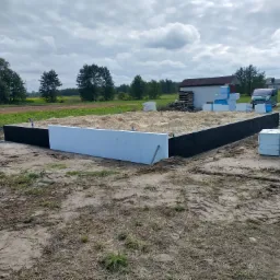 Fundament budynku w trakcie izolacji, widoczne czarne ściany fundamentowe oraz białe płyty izolacyjne, na ziemi leżą opakowania materiałów izolacyjnych, w tle budynek gospodarczy i zielony samochód...