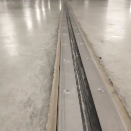 Szczelina dylatacyjna w posadzce betonowej parkingu podziemnego, zabezpieczona metalowymi profilami skręconymi śrubami.