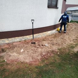 ABW Izolacje Wody Bartłomiej Wojewodzki - Prace ziemne przy fundamentach budynku jednorodzinnego, widoczna łopata wbita w ziemię, pracownik w odzieży roboczej i żółte wiadro.