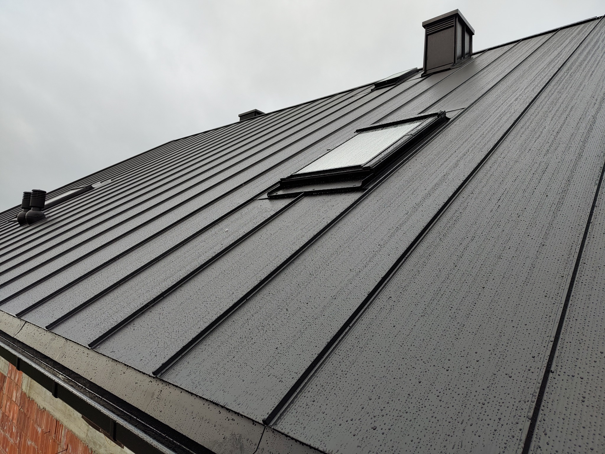 Nowoczesny, metalowy dach w kolorze grafitowym z oknem dachowym i kominem, pokryty kroplami deszczu w pochmurny dzień. Widoczny fragment muru z czerwonej cegły.