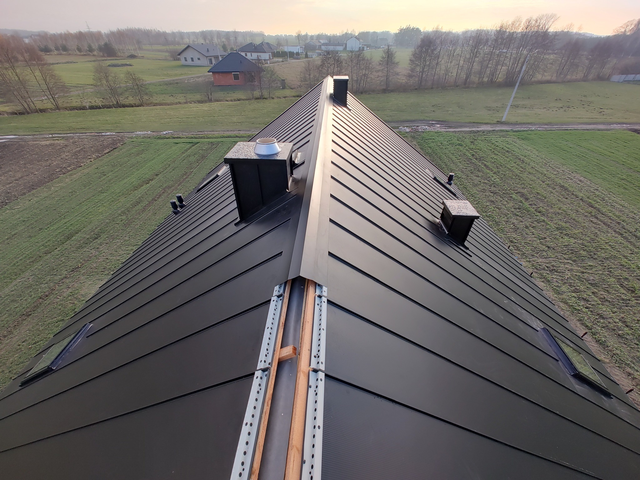 Czarny, blaszany dach dwuspadowy z kominami i oknami dachowymi w trakcie montażu, widok z góry na konstrukcję nośną.