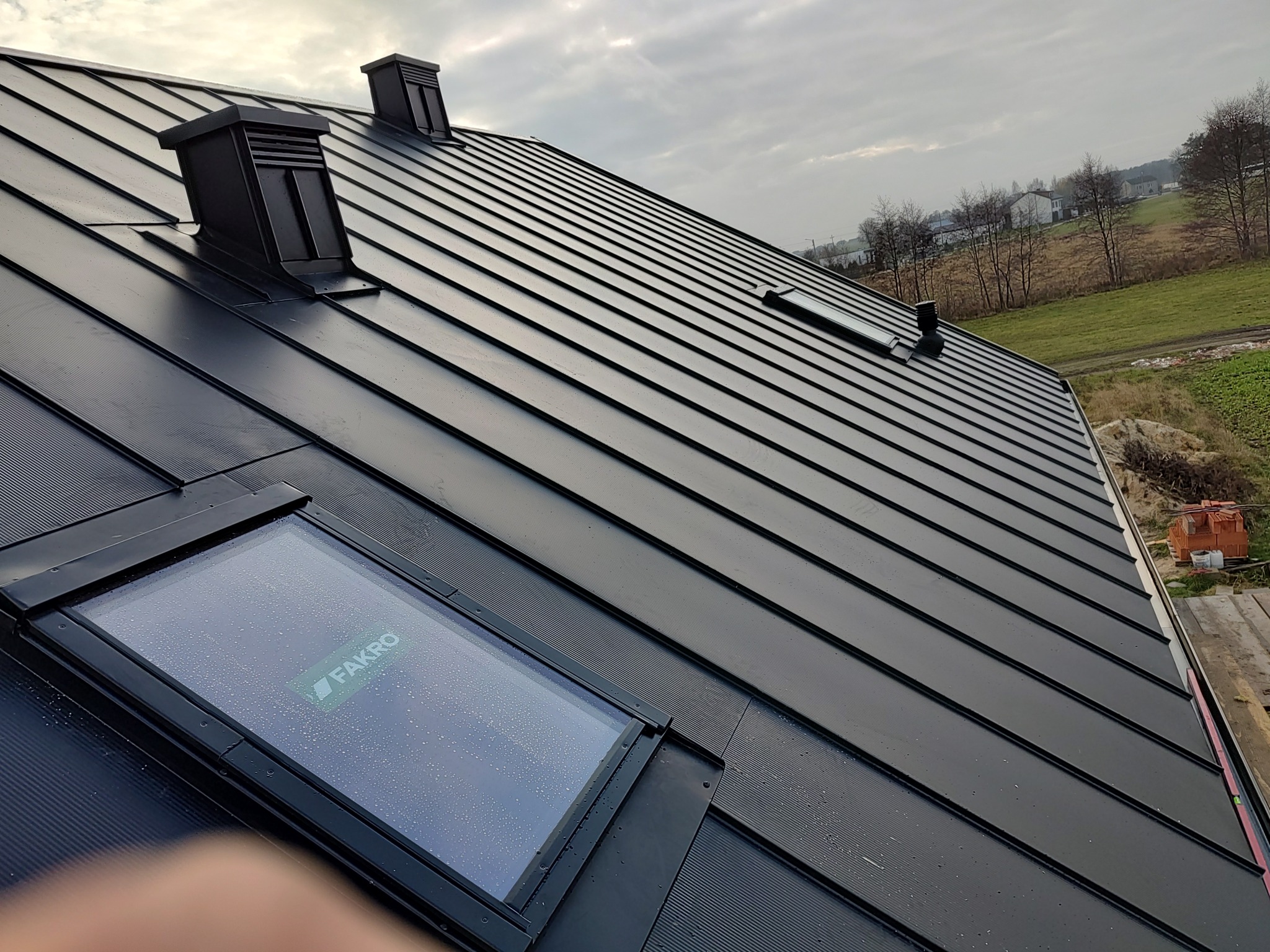 Czarny, metalowy dach z dwoma kominami i oknem dachowym marki Fakro, widok z dołu w Opocznie.