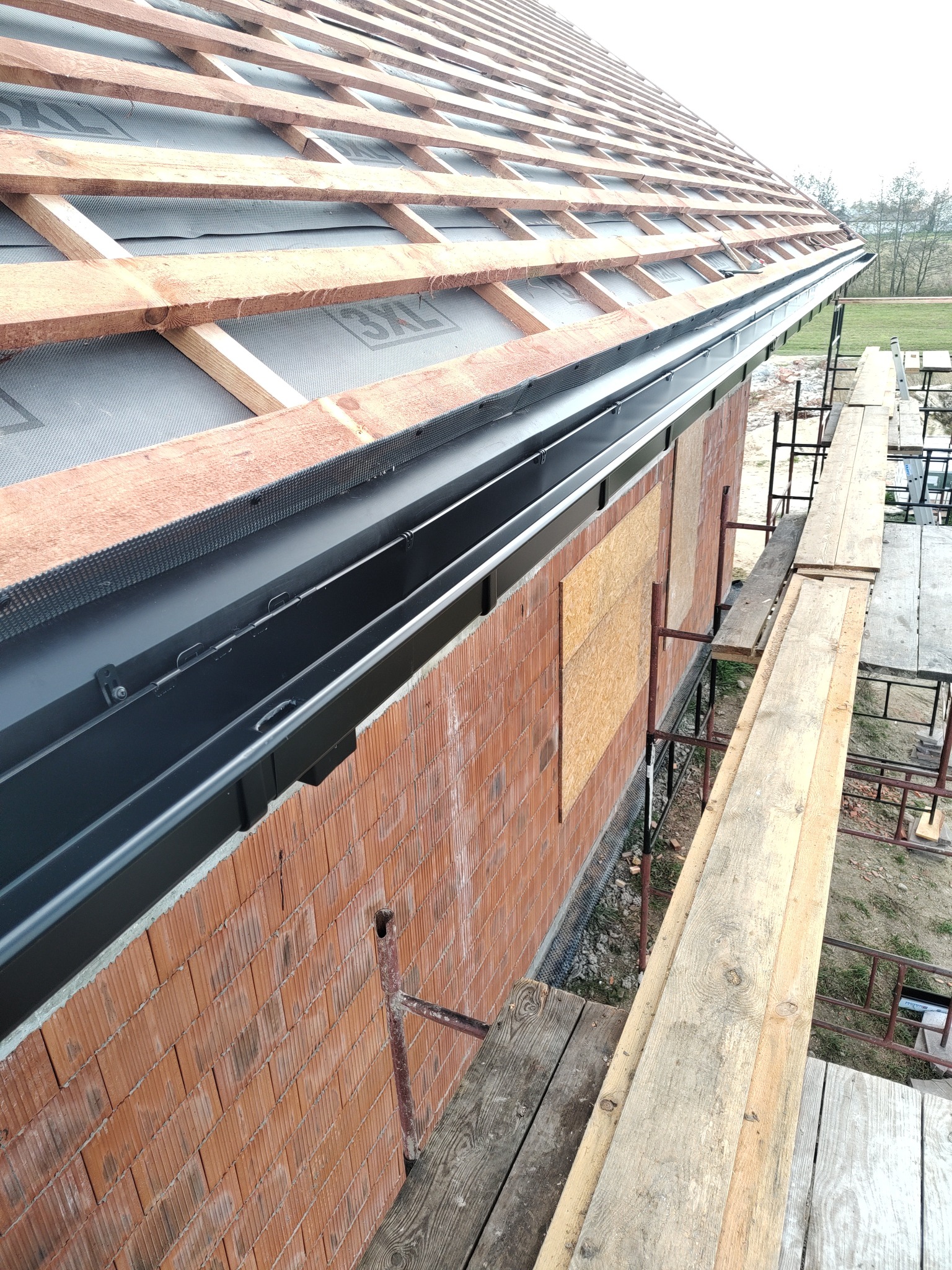 Montaż czarnych rynien na nowym budynku z cegły, widoczna konstrukcja dachu z membraną i rusztowania.
