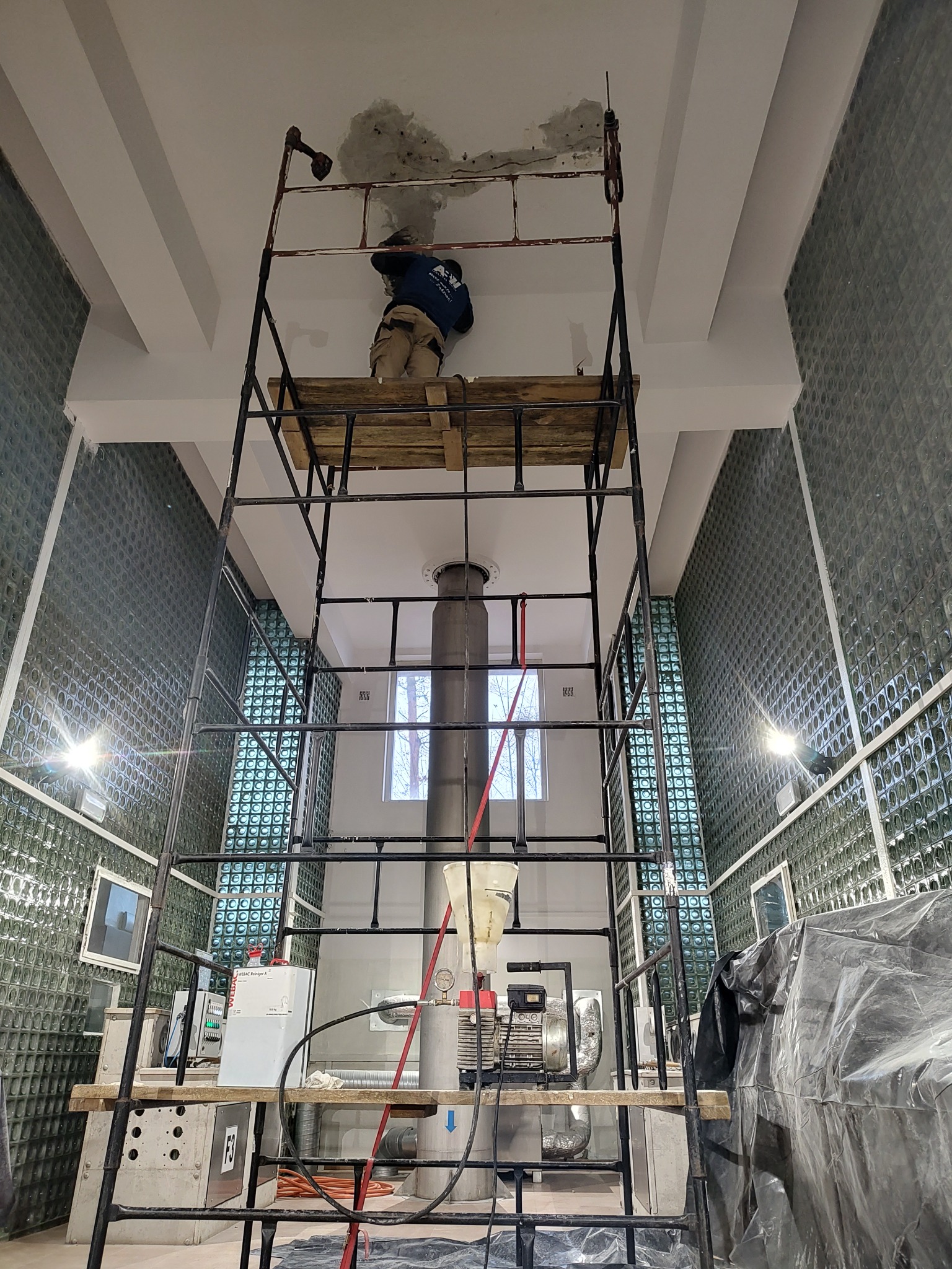 Pracownik na rusztowaniu naprawia uszkodzony sufit w pomieszczeniu z ścianami wyłożonymi szklanymi pustakami, widoczne maszyny i instalacje.