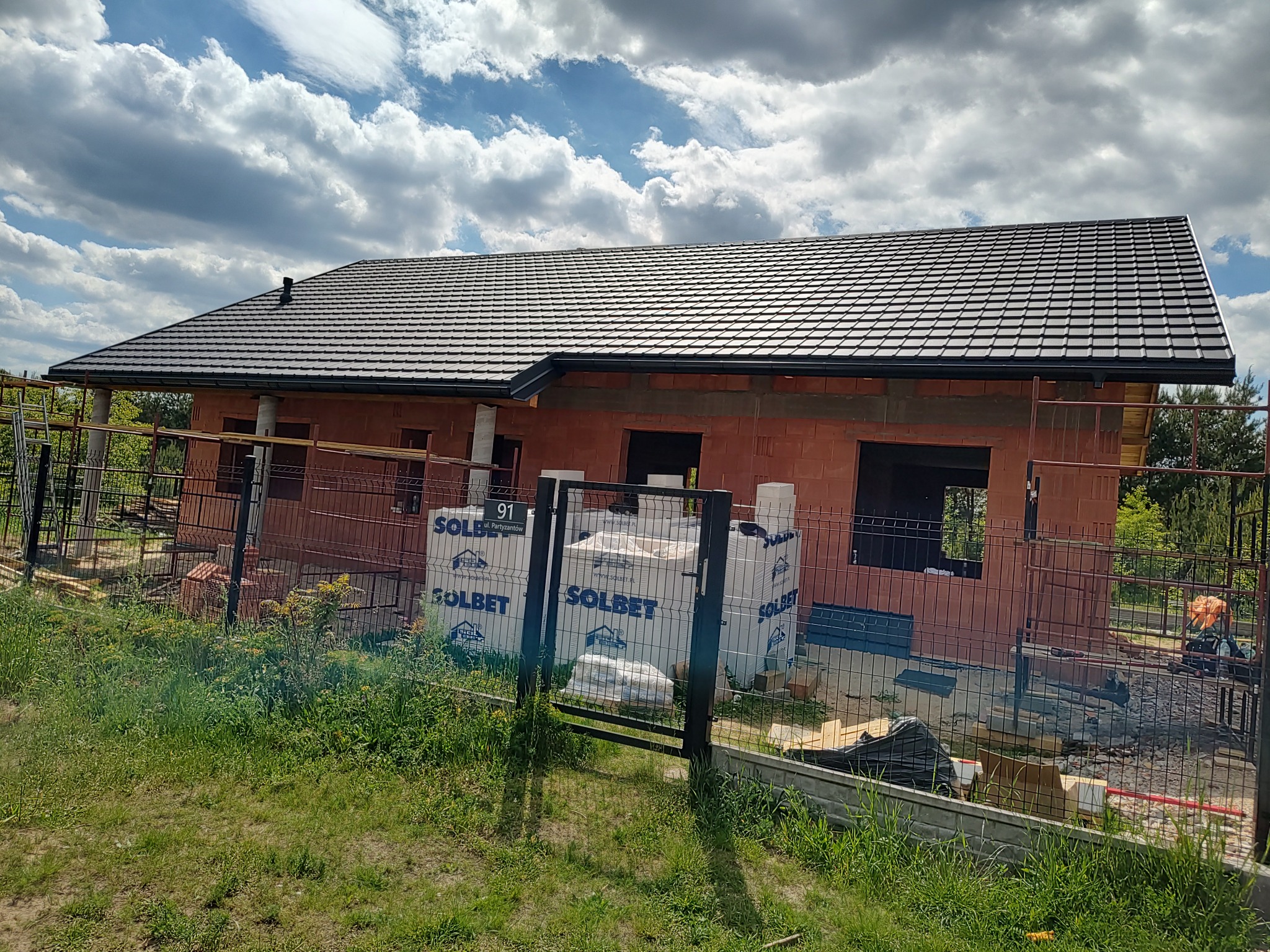 Dom w trakcie budowy z ciemnym dachem z dachówki, ogrodzony siatką. Widoczne pakiety materiałów budowlanych Solbet.