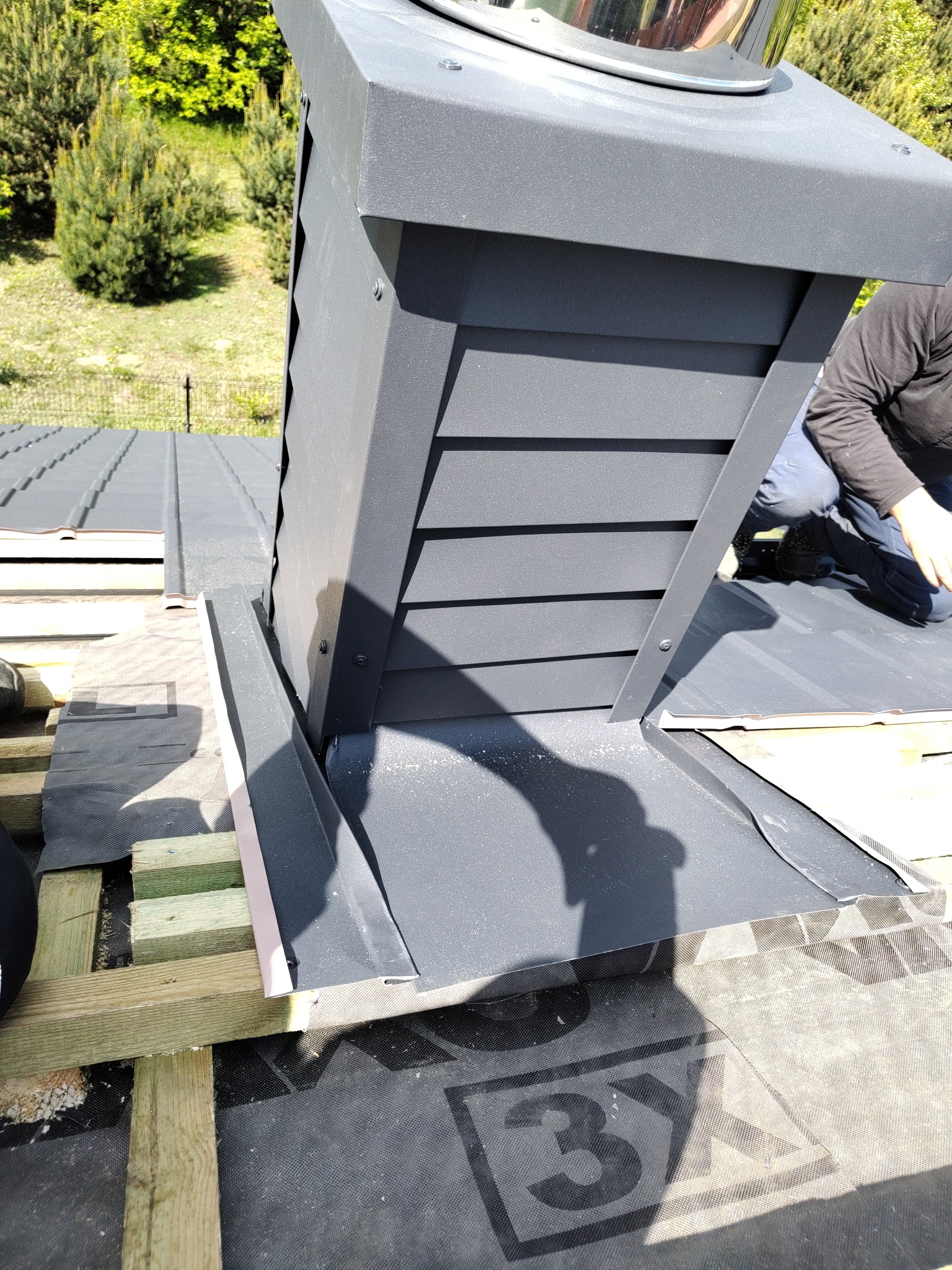 Montaż komina systemowego na dachu pokrytym blachą na rąbek stojący. Widoczne elementy konstrukcji dachu i membrana dachowa. Prace dekarskie w słoneczny dzień.