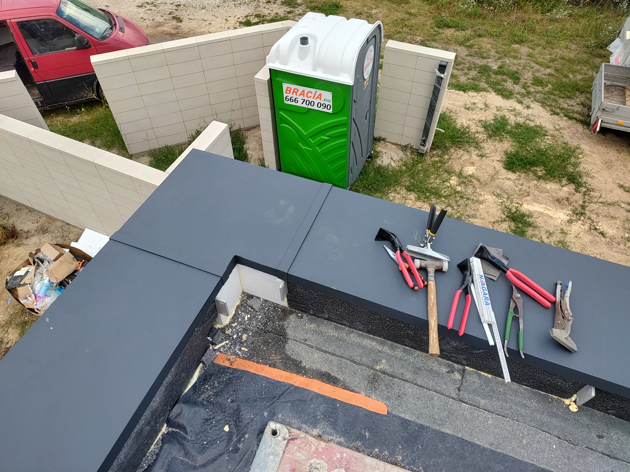 Nowoczesne krycie dachu z narzędziami dekarskimi na tle przenośnej toalety i częściowo widocznego samochodu dostawczego. Detal wykończenia.