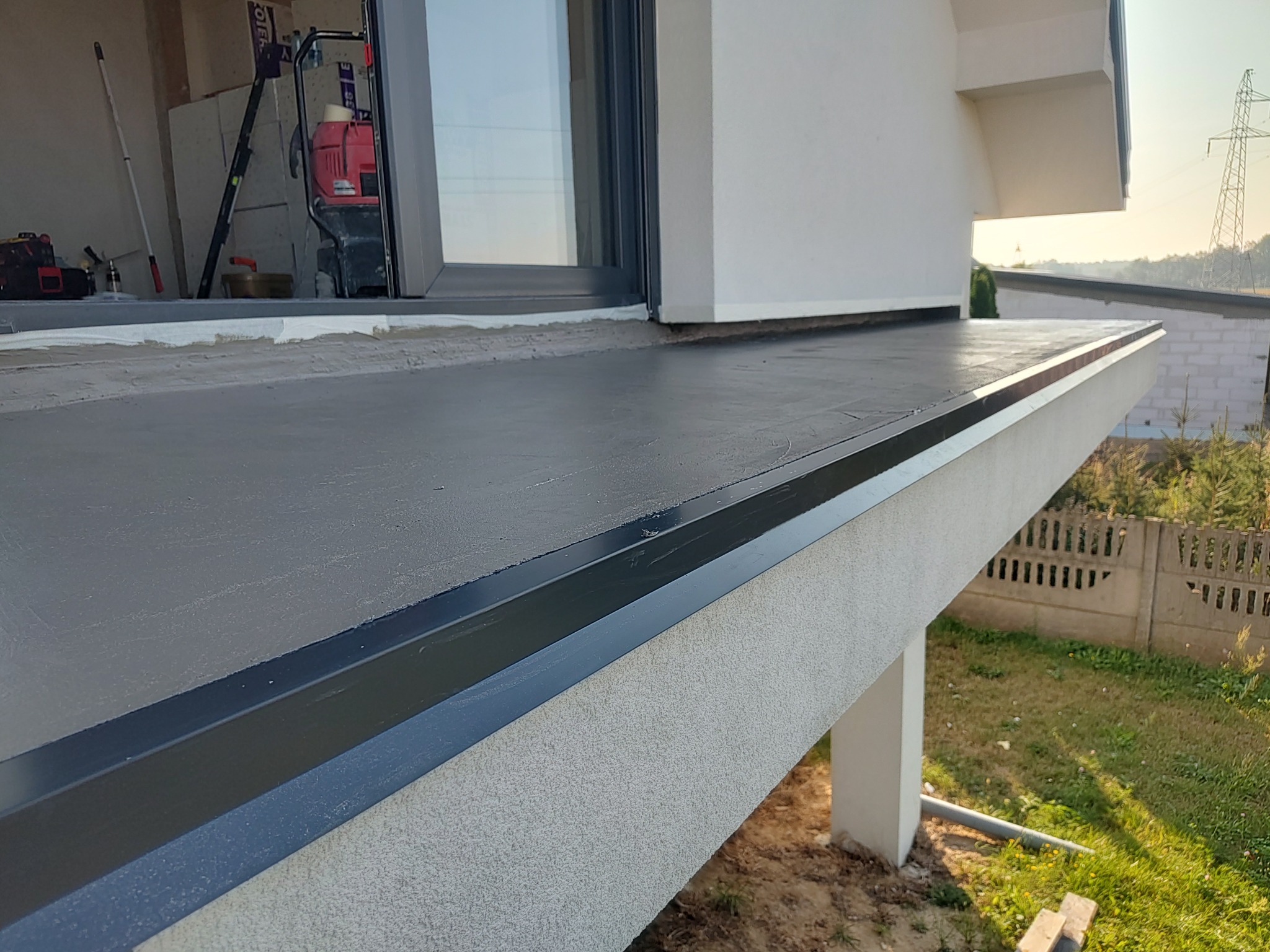 Szary, płaski dach po hydroizolacji, z widocznym oknem i fragmentem budynku. W tle zielony krajobraz i słup wysokiego napięcia. Widoczne narzędzia pracy.