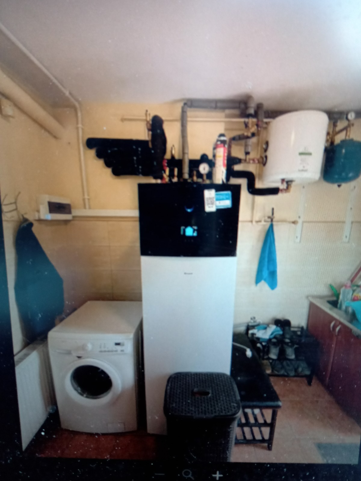 Biała pompa ciepła z czarnym panelem górnym, zainstalowana w pomieszczeniu gospodarczym obok pralki i bojlera, z widocznymi rurami instalacyjnymi pod sufitem i niebieskim ręcznikiem na ścianie.