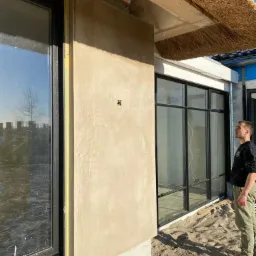 Zewnętrzna ściana budynku z nałożonym tynkiem o nierównej fakturze, widoczny fragment okna z czarną ramą, fragment strzechy i pracownik w tle.