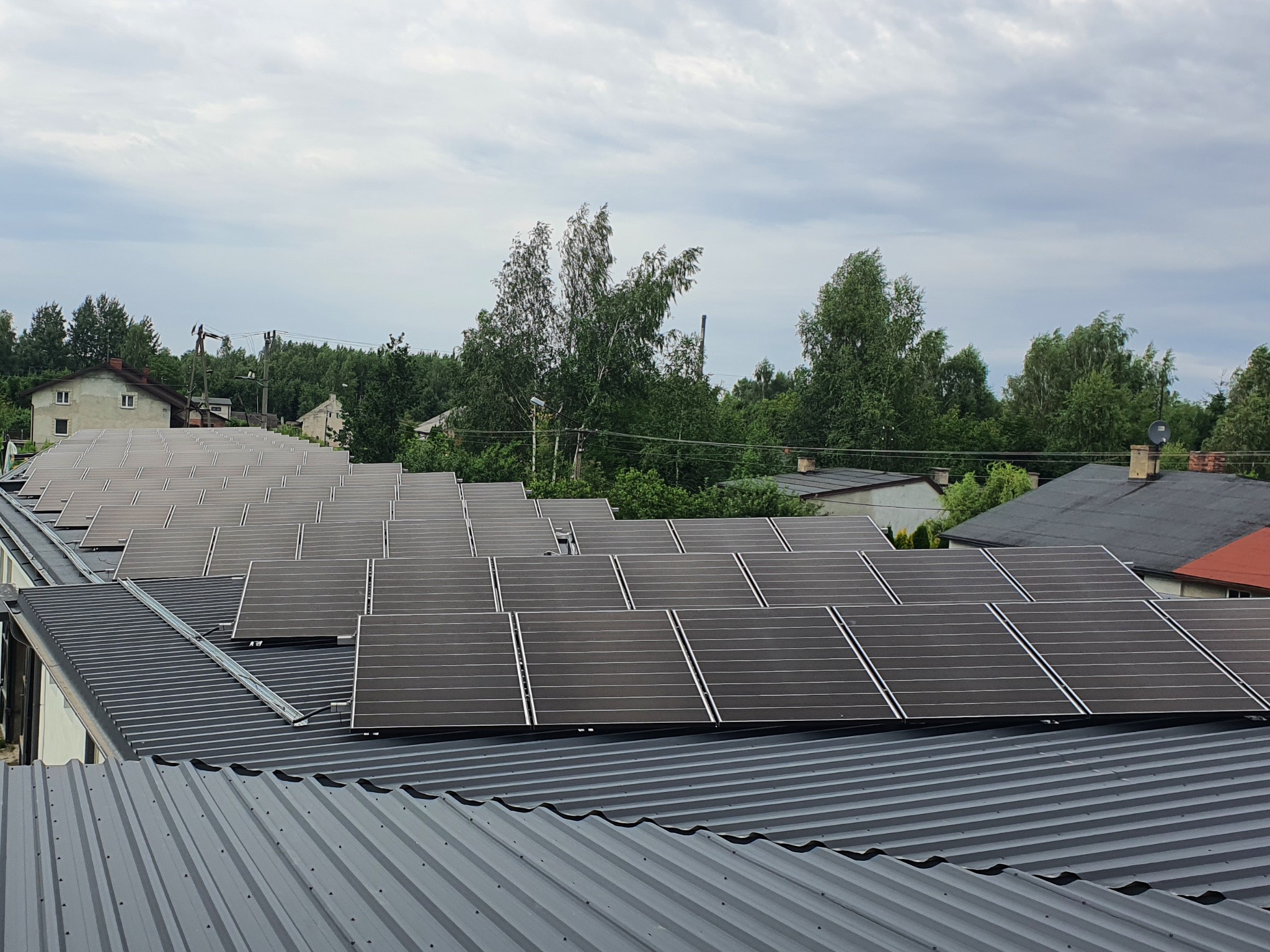 Panele fotowoltaiczne zamontowane na falistej blasze dachowej, widok z góry, otoczone zielenią drzew i zabudowaniami mieszkalnymi w tle.