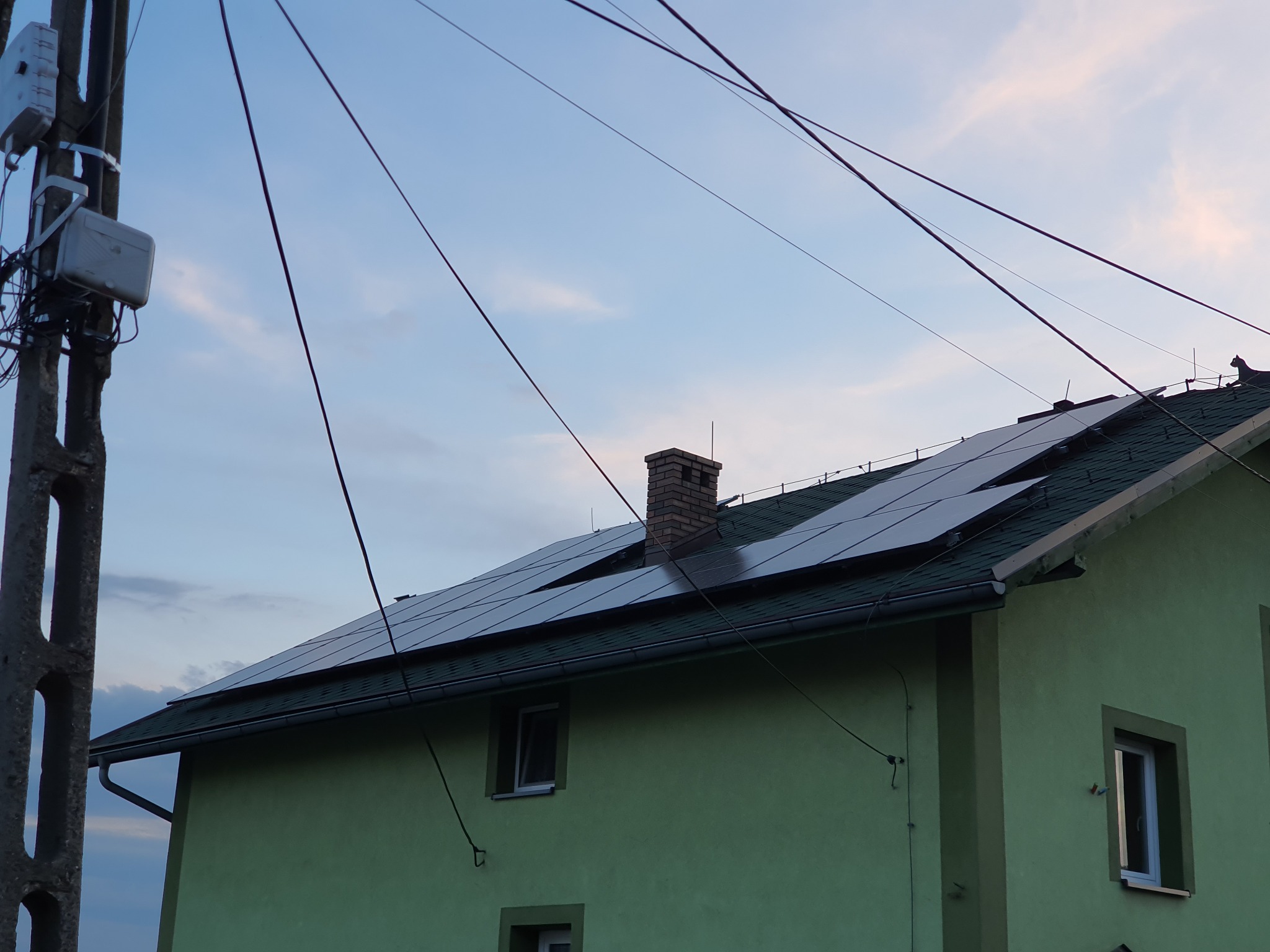 Panele słoneczne na dachu zielonego domu z ceglanym kominem, widoczne słupy i przewody elektryczne na tle pochmurnego nieba.