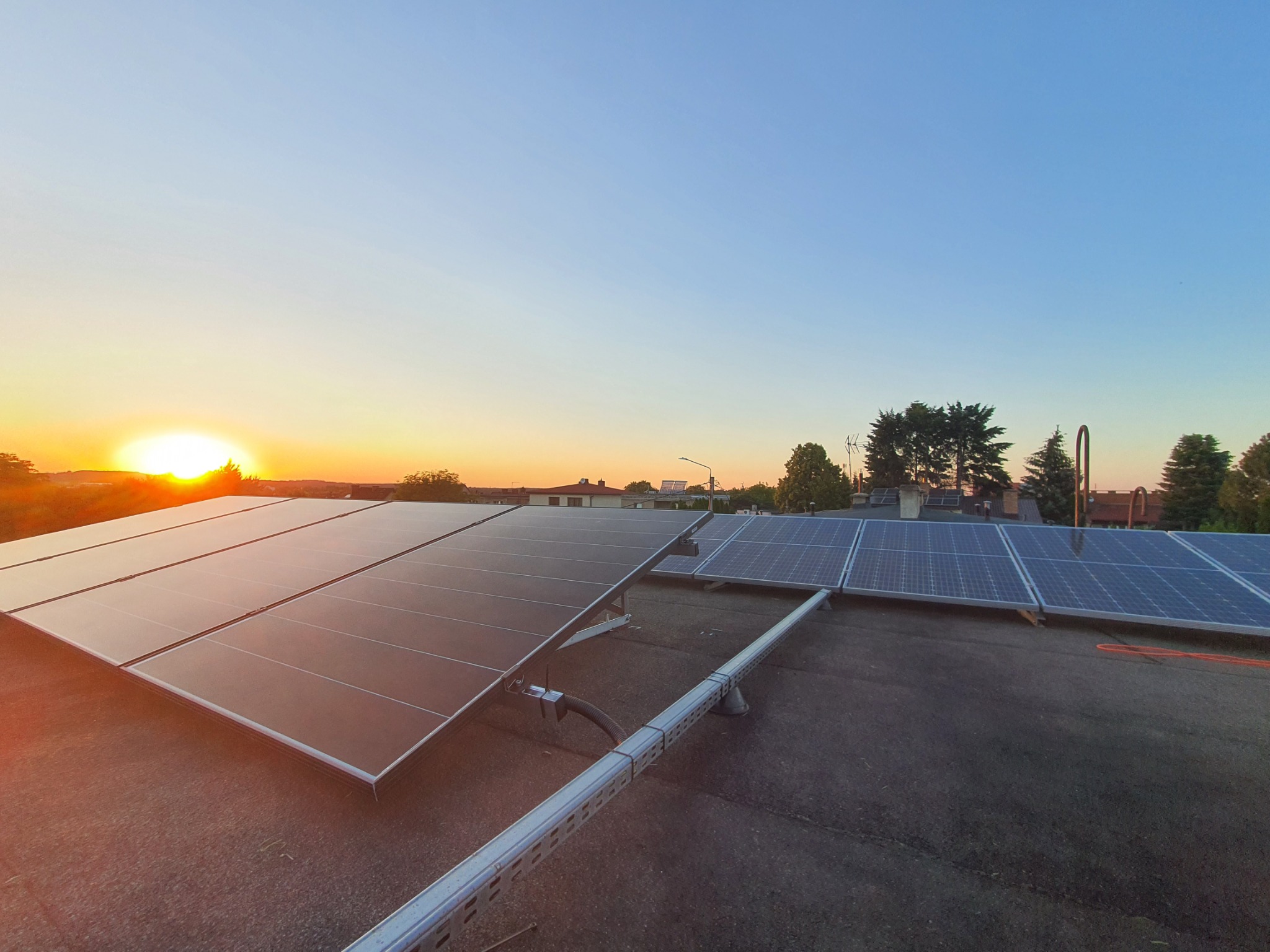 Panele słoneczne na dachu budynku oświetlone promieniami zachodzącego słońca, widok na okoliczne domy i drzewa