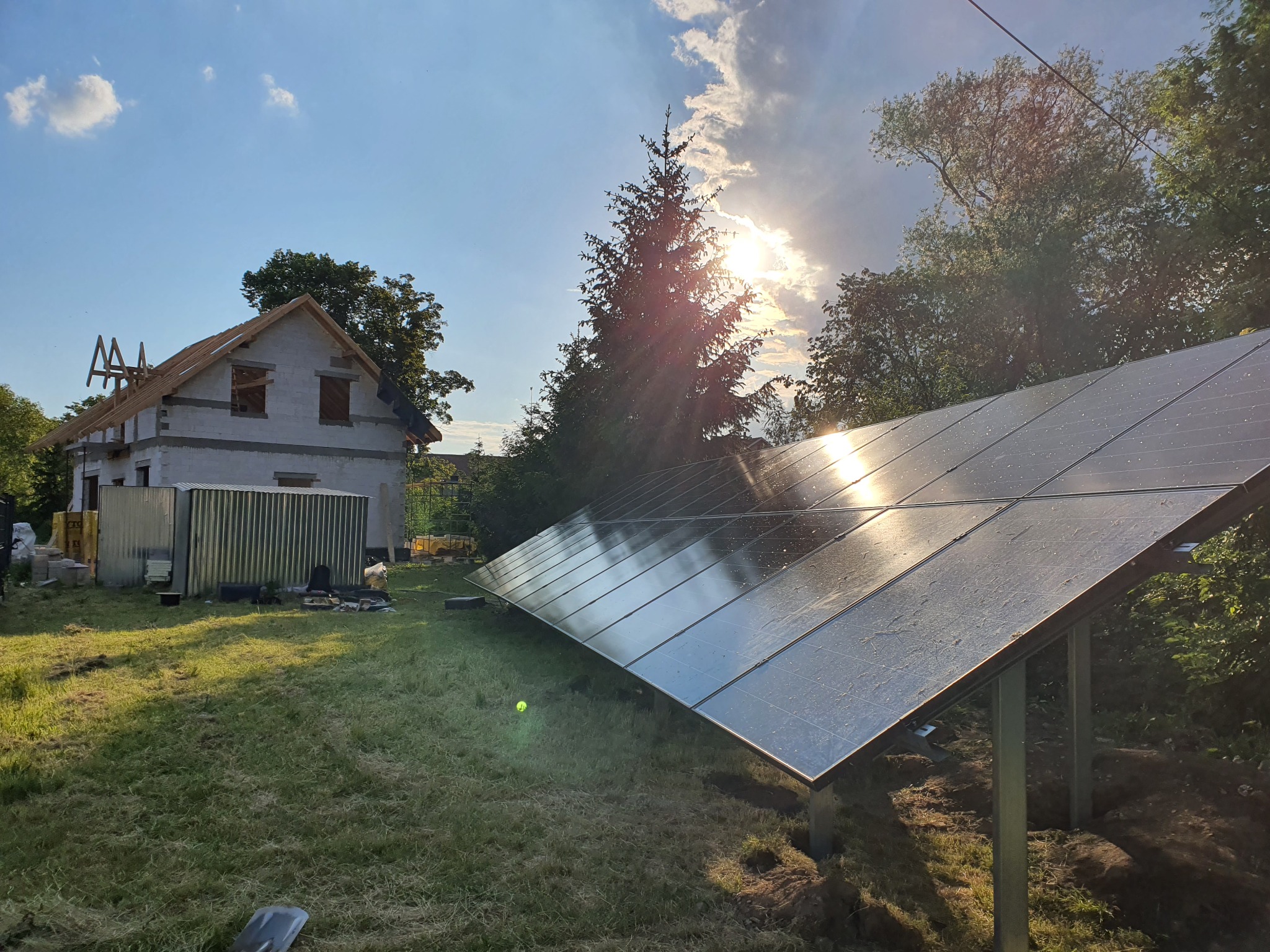Panele słoneczne zamontowane na metalowych wspornikach na trawiastym terenie, w tle dom w trakcie budowy i drzewa, oświetlone popołudniowym słońcem.