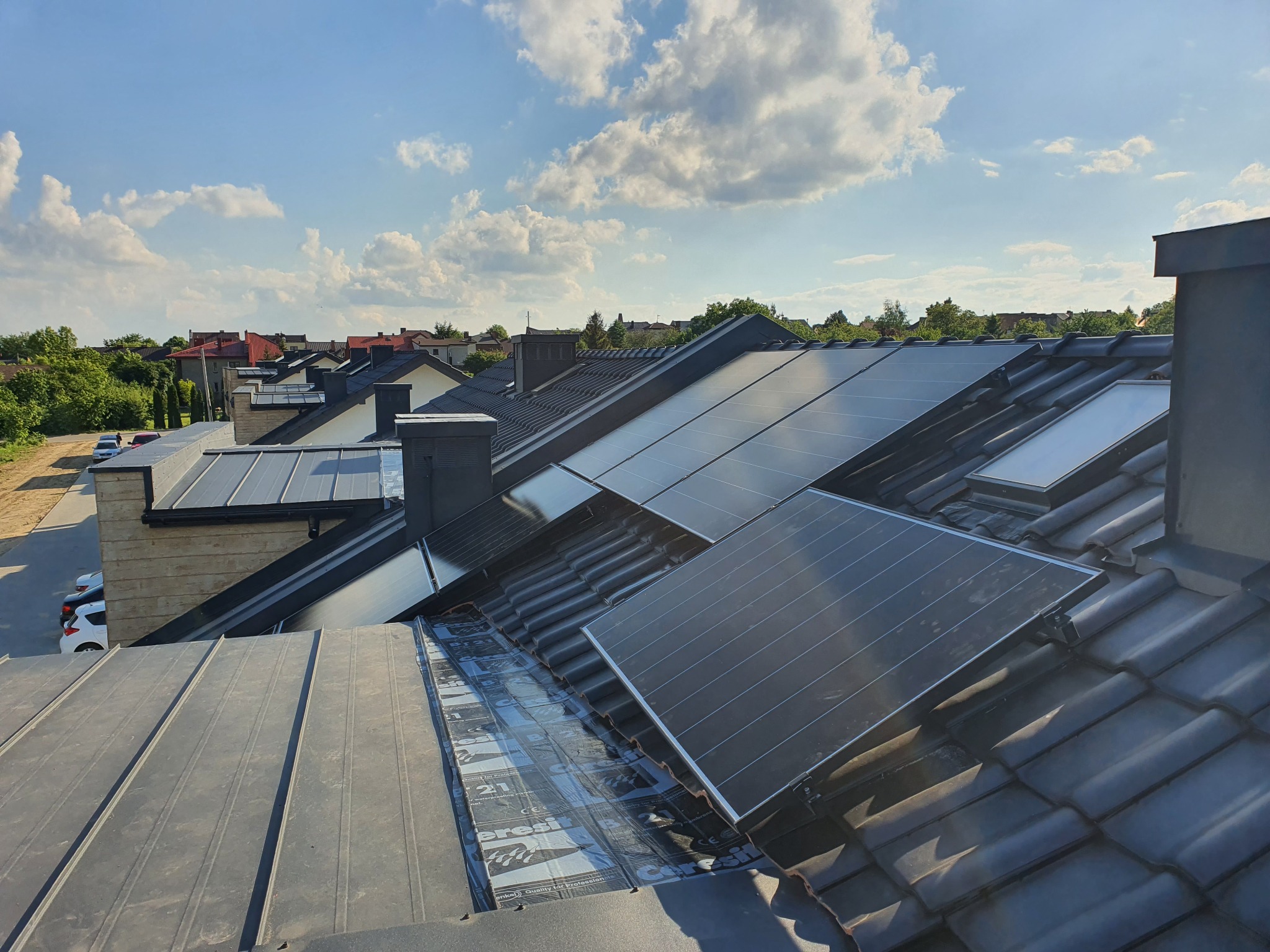 Panele słoneczne zamontowane na dachu z ciemnej dachówki, widoczne membrany hydroizolacyjne, w tle zabudowa jednorodzinna i błękitne niebo z chmurami.