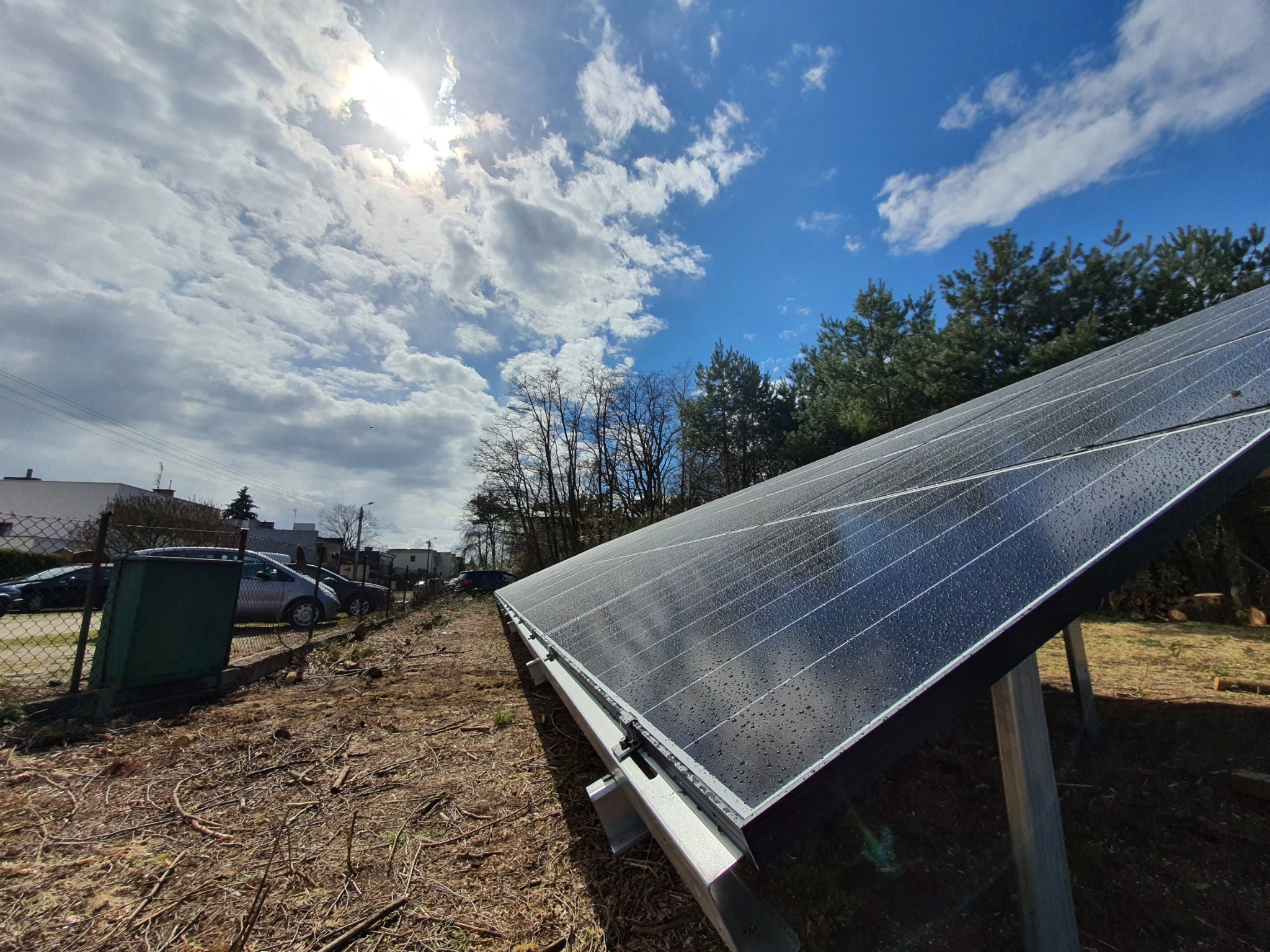 Panel słoneczny z kroplami deszczu na powierzchni, widoczny na tle błękitnego nieba z chmurami i drzewami. Montaż na gruncie z widocznymi elementami konstrukcyjnymi.