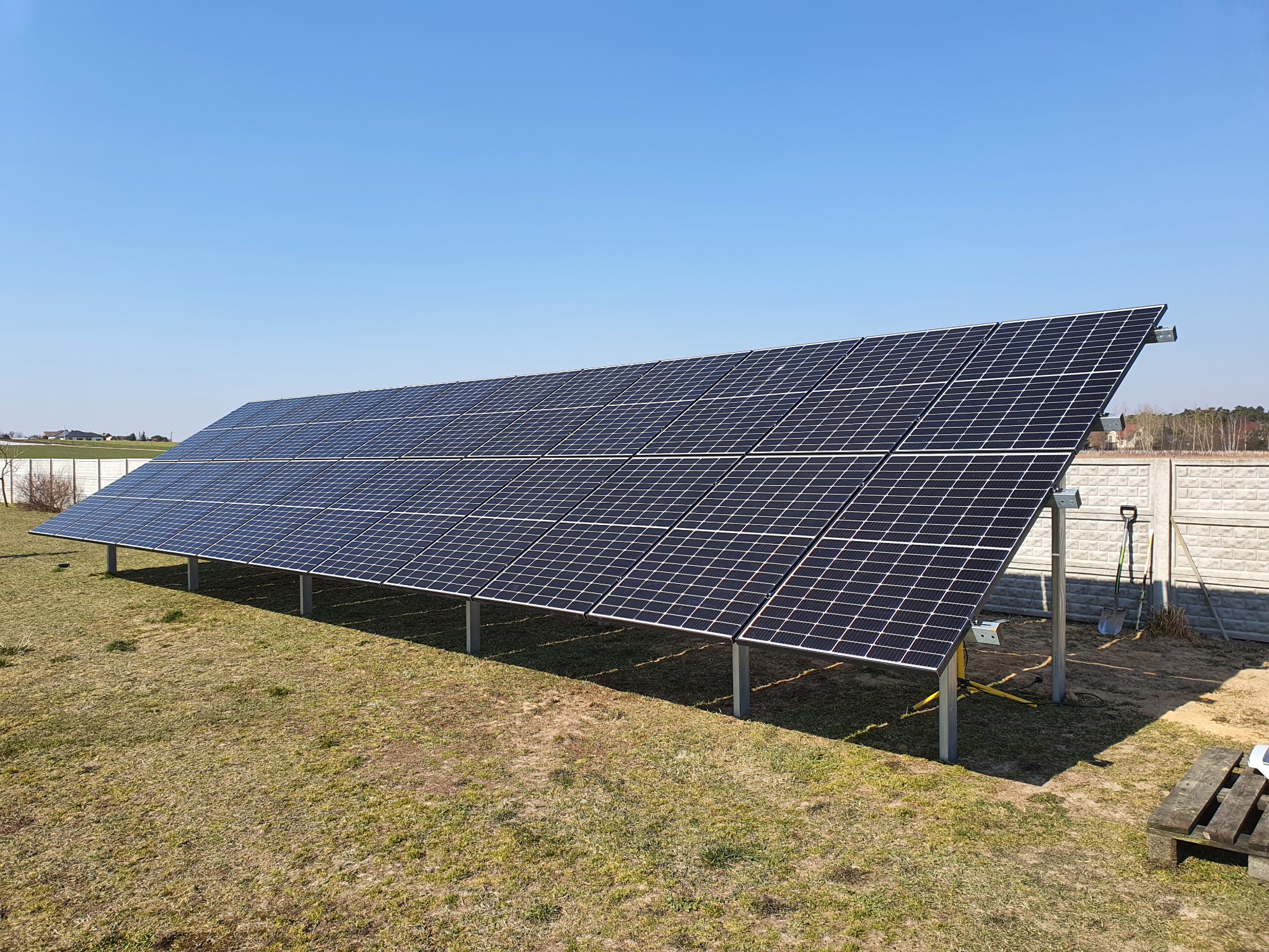 Rozległa instalacja paneli słonecznych na niskim stelażu, z widocznymi narzędziami i paletą w tle, na trawiastym terenie pod błękitnym niebem.