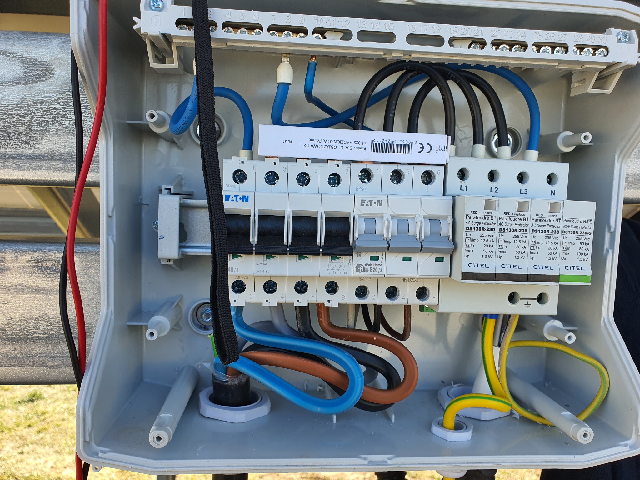 Otwarta skrzynka elektryczna z widocznymi bezpiecznikami Eaton, ogranicznikami przepięć Citel i przewodami w różnych kolorach, zamocowana na zewnątrz budynku.