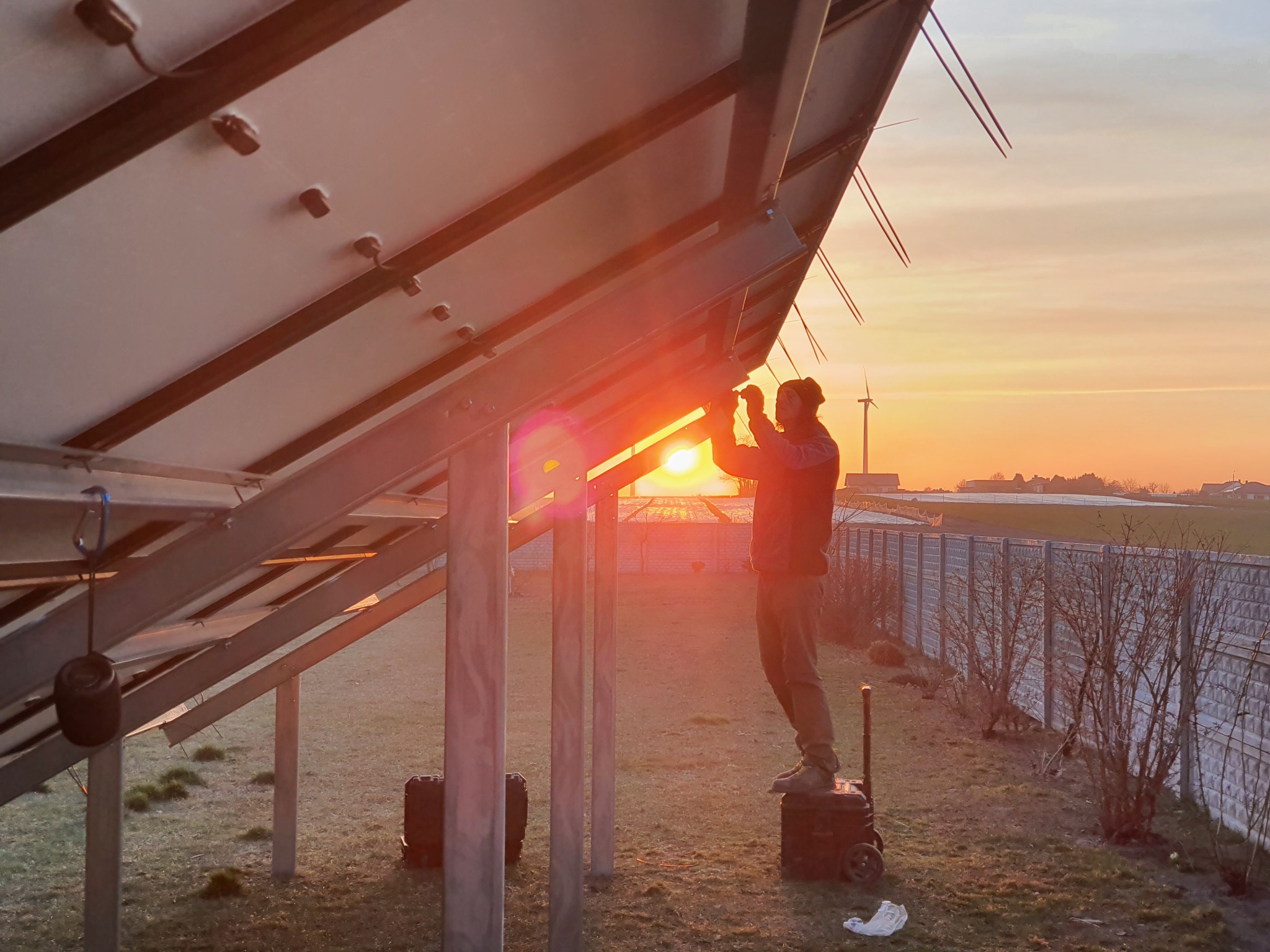 Instalator na podwyższeniu, montuje panele słoneczne na konstrukcji stalowej, w tle zachód słońca i turbina wiatrowa.