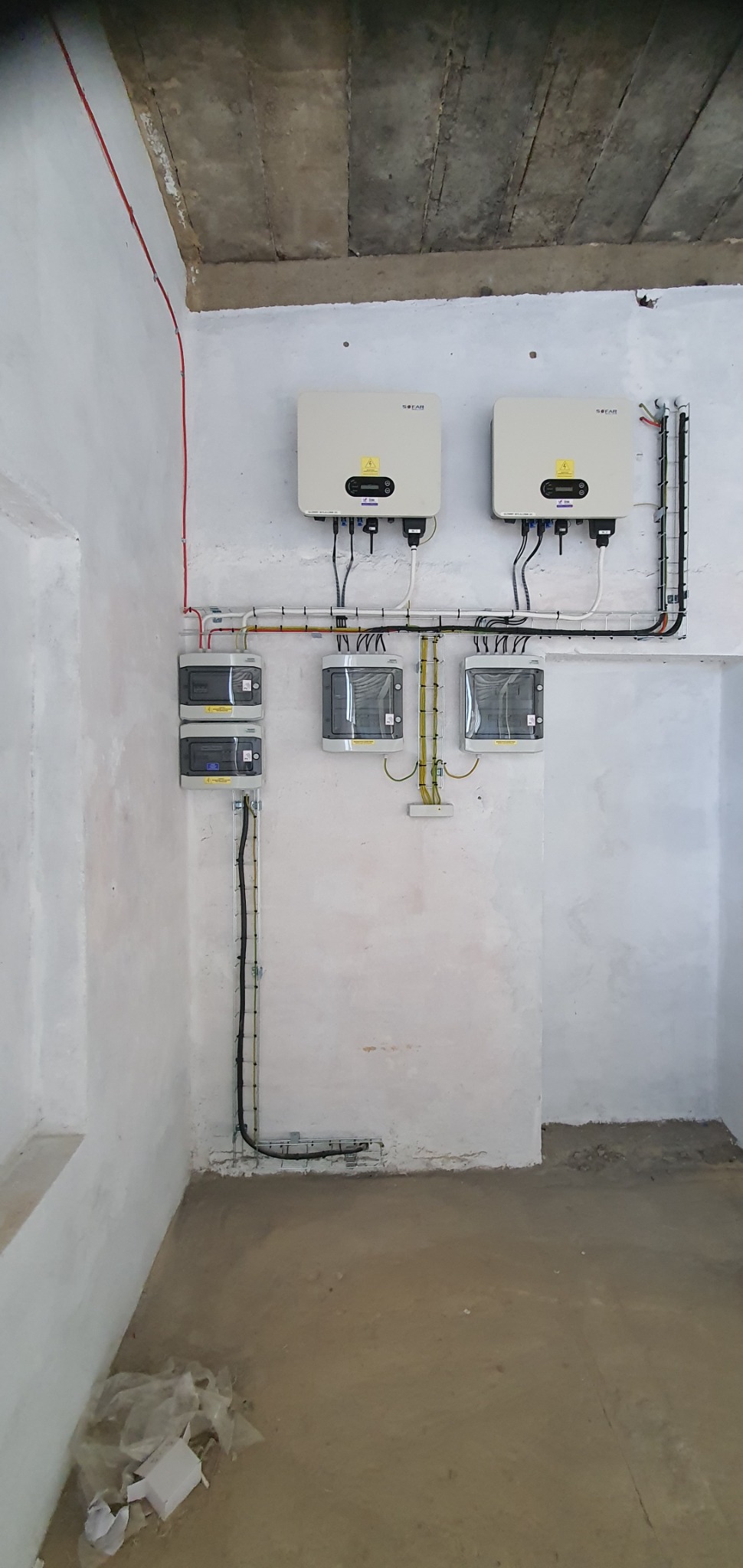 Dwa białe inwertery fotowoltaiczne marki SOFAR zamontowane na ścianie w surowym pomieszczeniu, z widocznymi przewodami i zabezpieczeniami elektrycznymi. Instalacja w trakcie realizacji, widoczne...