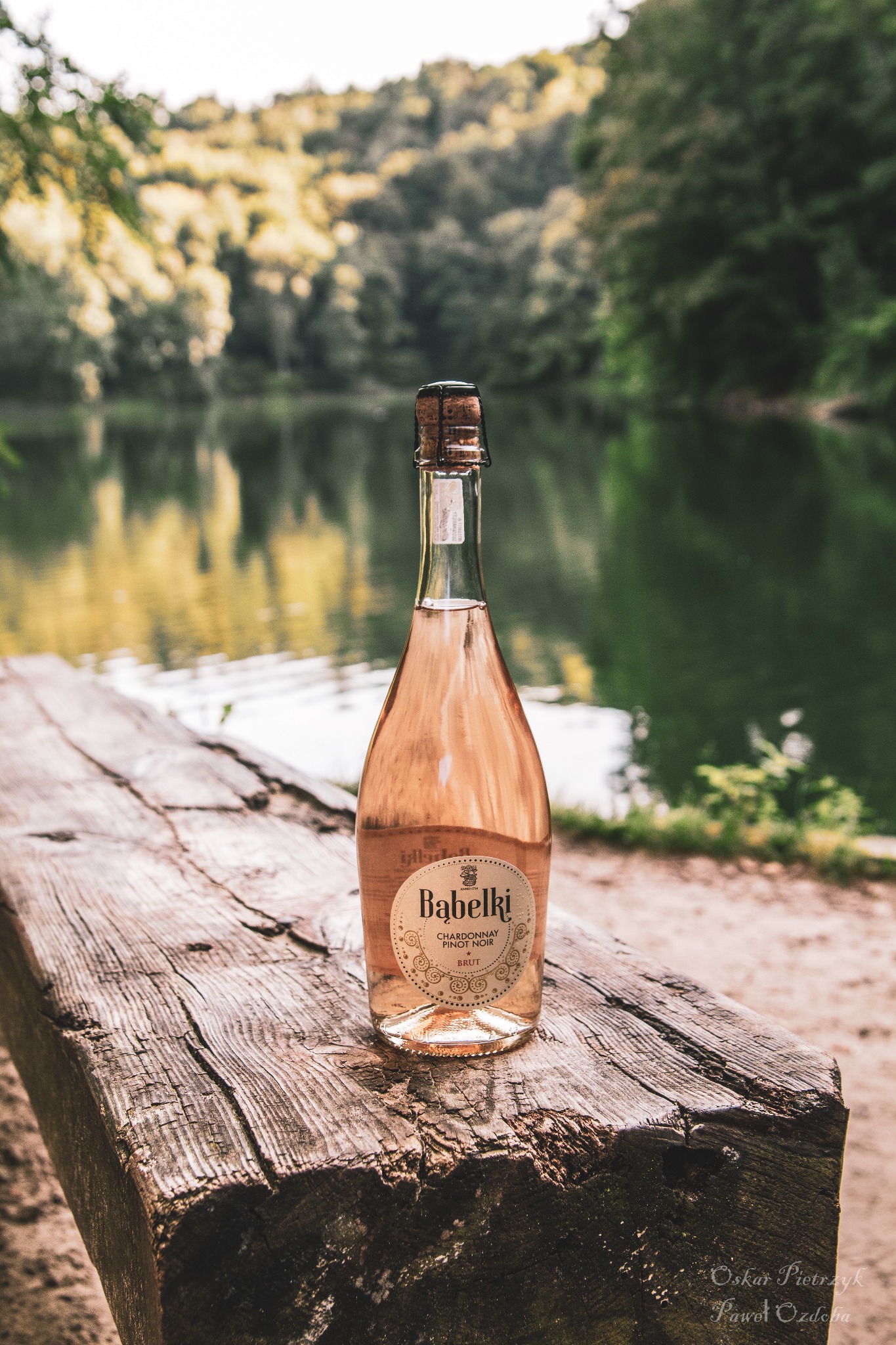 Butelka różowego wina musującego Babelki Chardonnay Pinot Noir Brut na surowym drewnianym stole z widokiem na jezioro i las w tle.