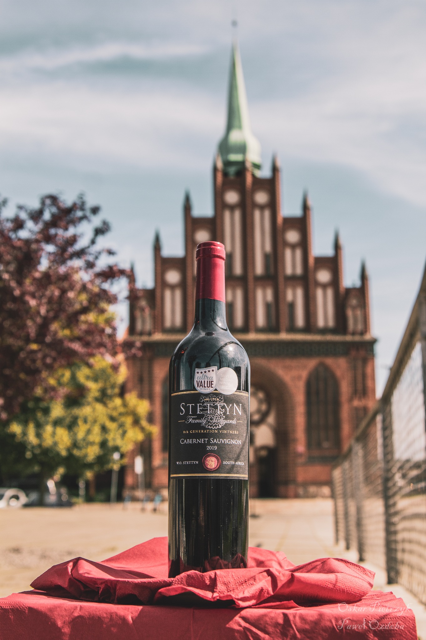 Butelka czerwonego wina Cabernet Sauvignon marki Stettyn na czerwonej tkaninie, na tle ceglanego kościoła z zieloną wieżą i drzewami.