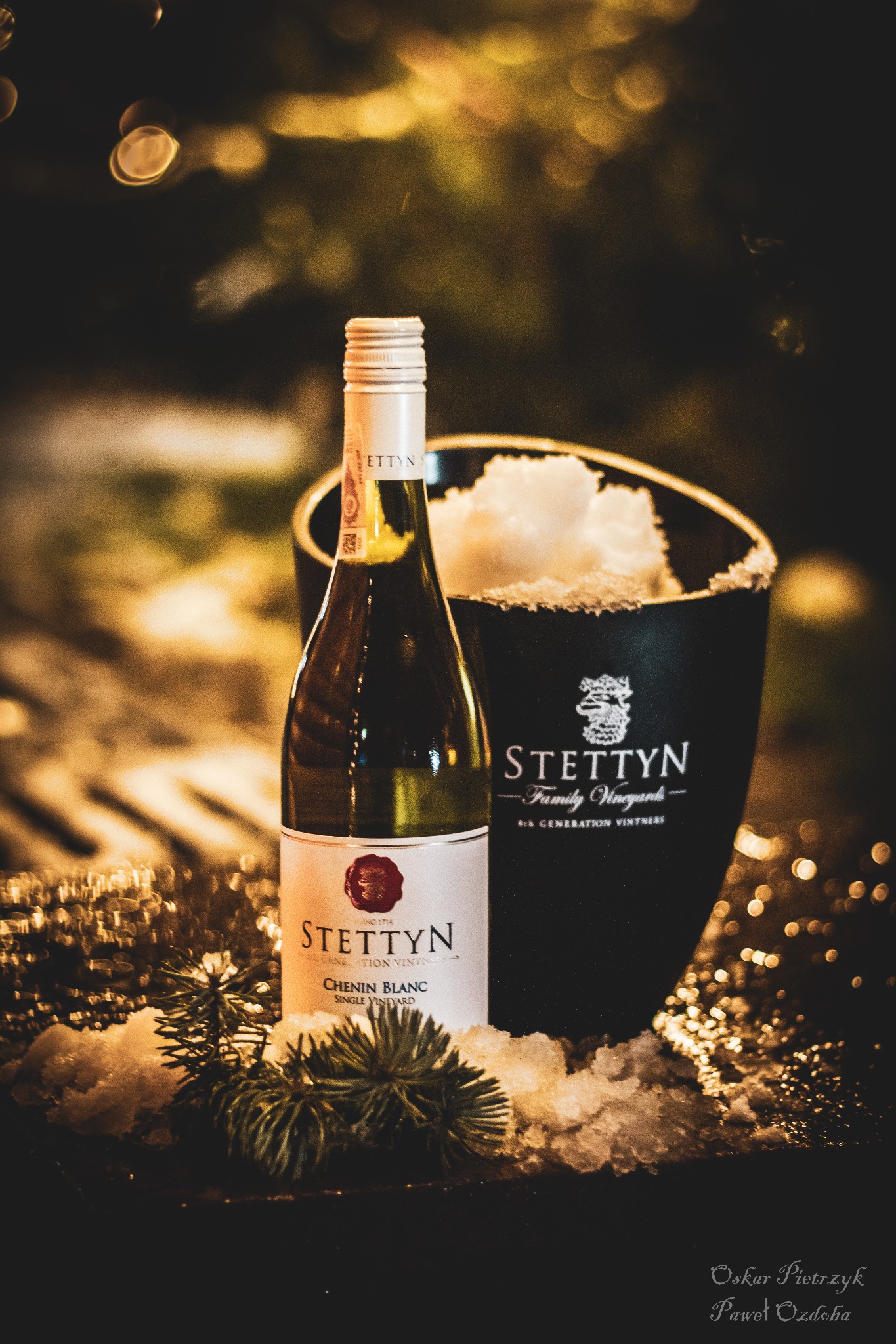 Butelka białego wina marki Stettyn Chenin Blanc w wiaderku z lodem, udekorowana gałązkami świerku, prezentowana w Szczecinie na ciemnym tle z rozmytymi światłami.