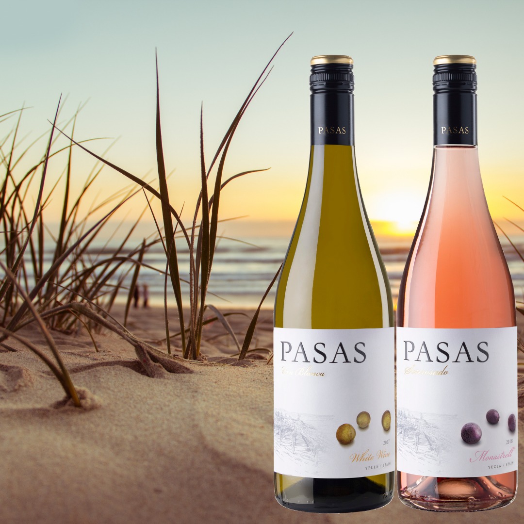 Dwie butelki wina, białe i różowe, na tle piaszczystej plaży o zachodzie słońca, z trawami na pierwszym planie.