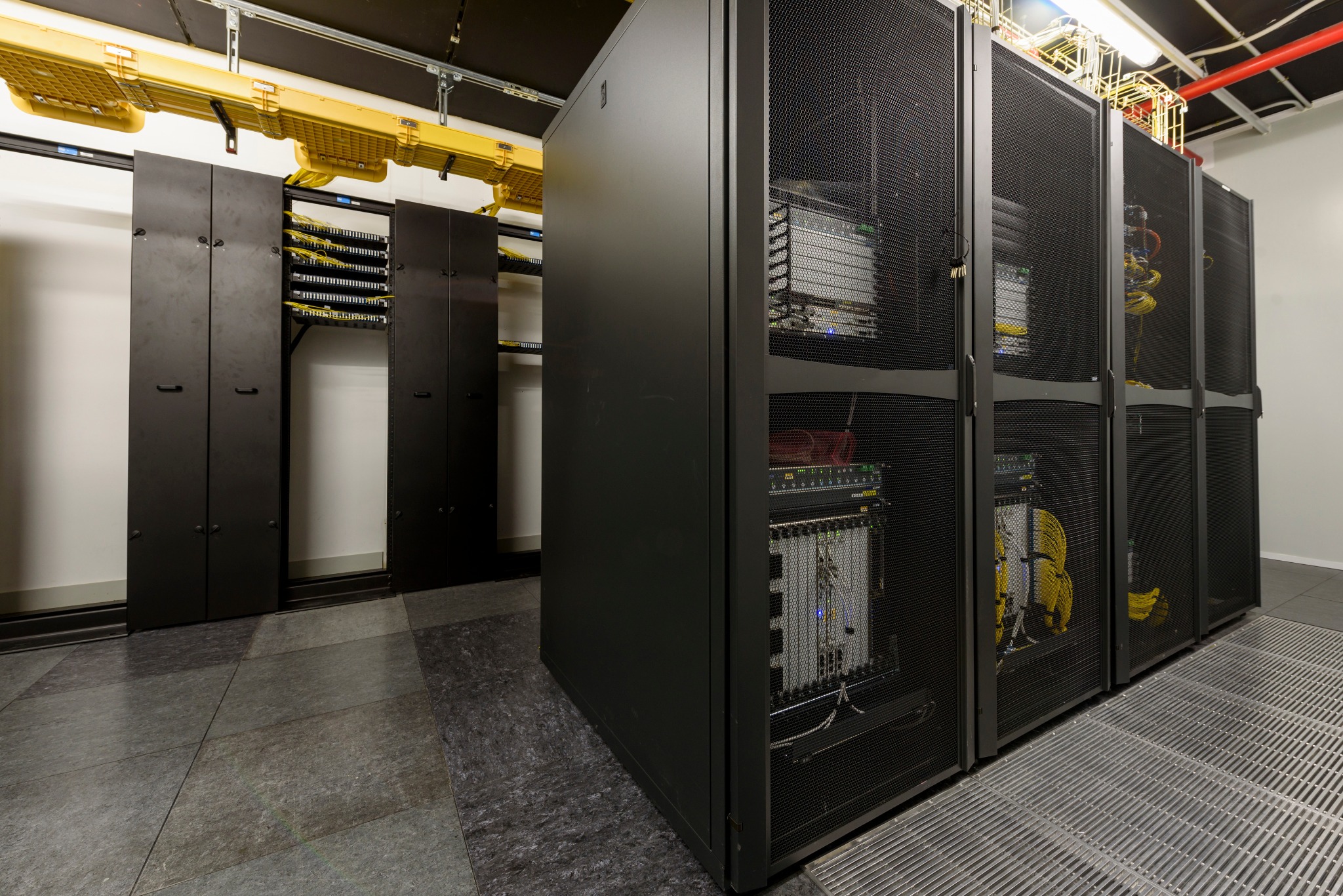 Rząd czarnych szaf serwerowych z widocznymi panelami krosowniczymi i okablowaniem w profesjonalnej serwerowni z podniesioną podłogą i systemem prowadzenia kabli pod sufitem.