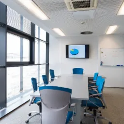 Nowoczesna sala konferencyjna z dużym oknem, białym stołem, niebieskimi krzesłami i ekranem wyświetlającym wykres kołowy.