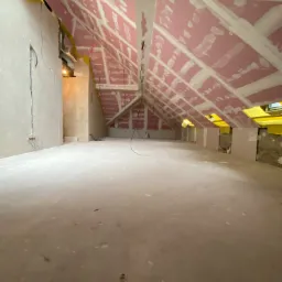 Poddasze w trakcie prac wykończeniowych: różowe płyty na skośnym suficie ze szpachlowanymi łączeniami, surowe ściany i podłoga, widoczne kable elektryczne.