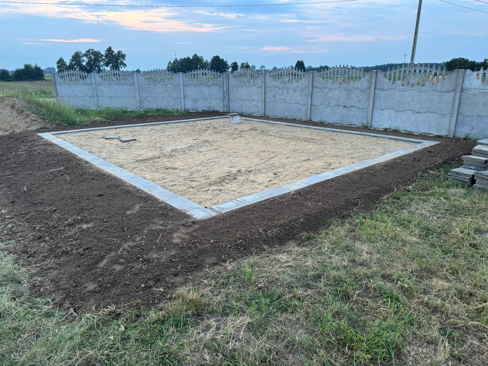 Przygotowany teren pod budowę, obramowany kostką brukową, wypełniony piaskiem, widoczny betonowy płot w tle.