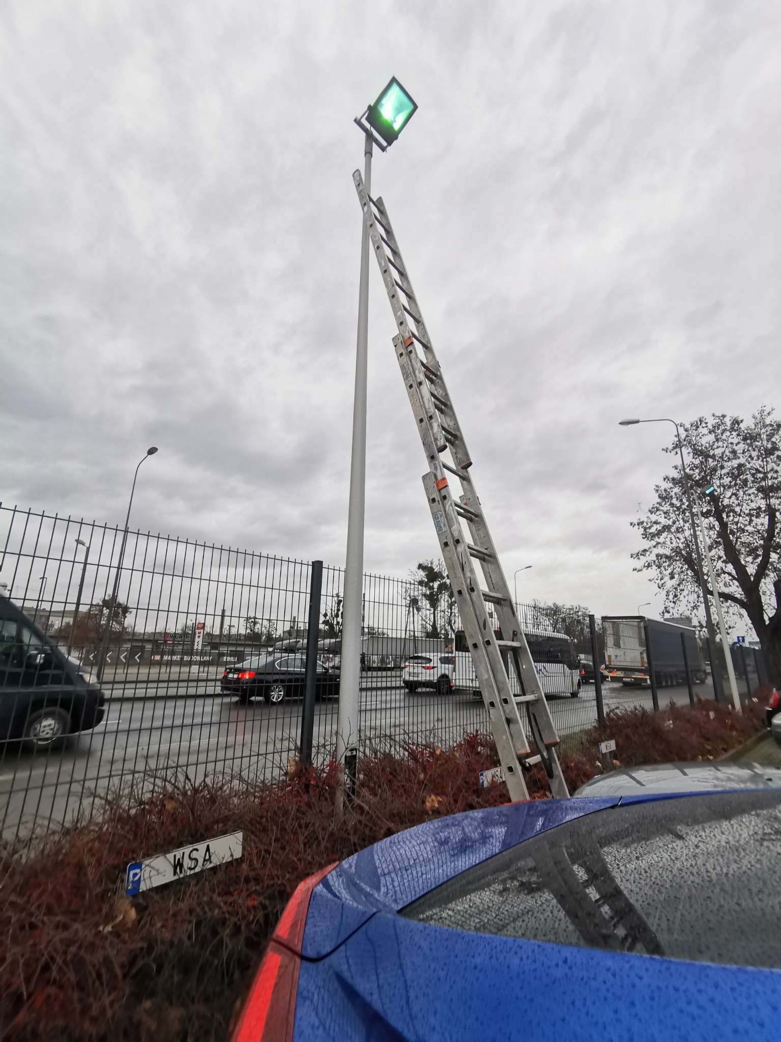 Metalowa drabina oparta o słup oświetleniowy z zielonym światłem na tle pochmurnego nieba, widok z parkingu z fragmentem niebieskiego samochodu w kadrze.